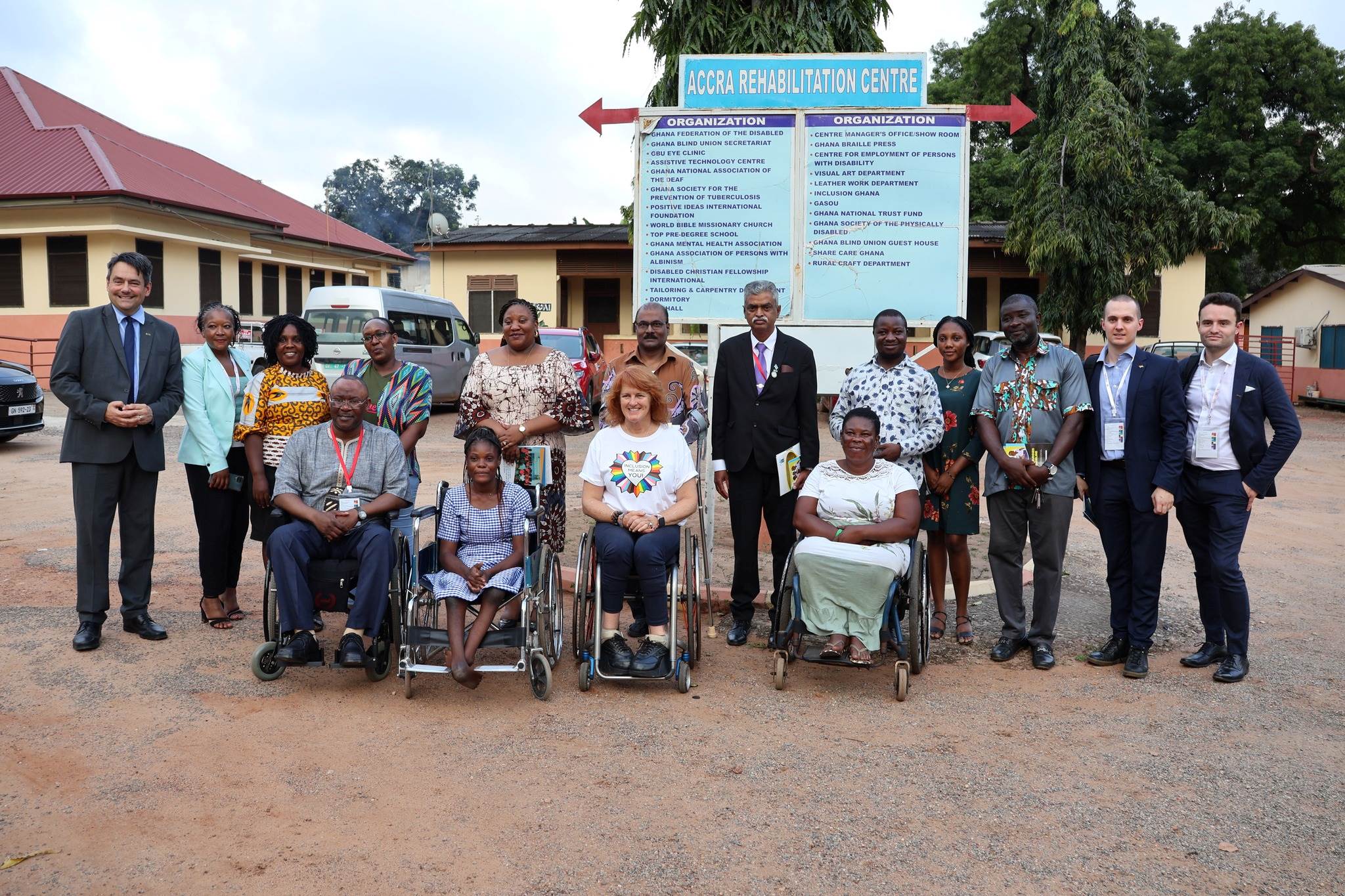 A group of people, including several wheelchair users, smile at the camera in front a sign for the Accra Rehabilitation Centre.